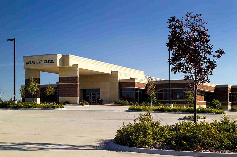 Exterior photo of the Hubbell‑built Wolfe Eye Clinic with its brick and stone exterior, covered main entrance, landscaped shrubs, and clear blue sky backdrop.