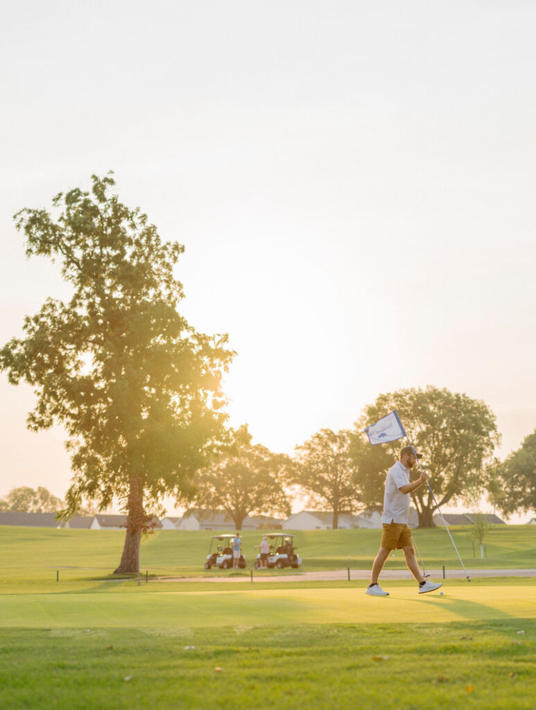 A person walks across a golf green holding a flag during sunset, with trees, golf carts, and a bright sky in the background.