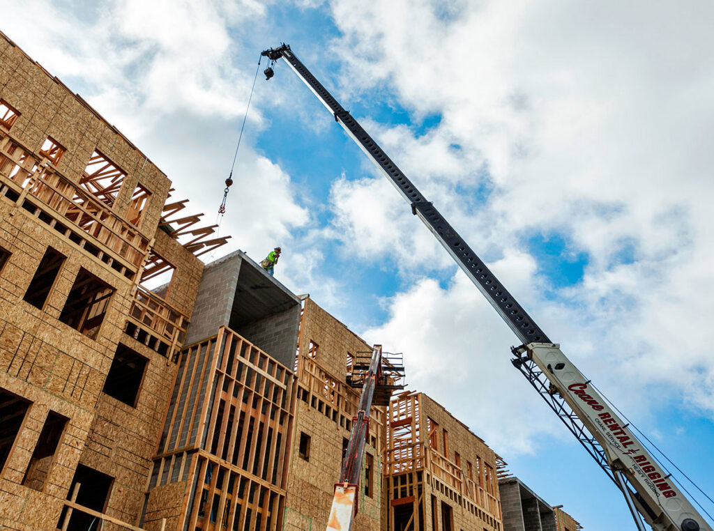 Construction workers assemble the upper levels of a multi‑story building framed in wood, while a large crane lifts materials against a partly cloudy sky.