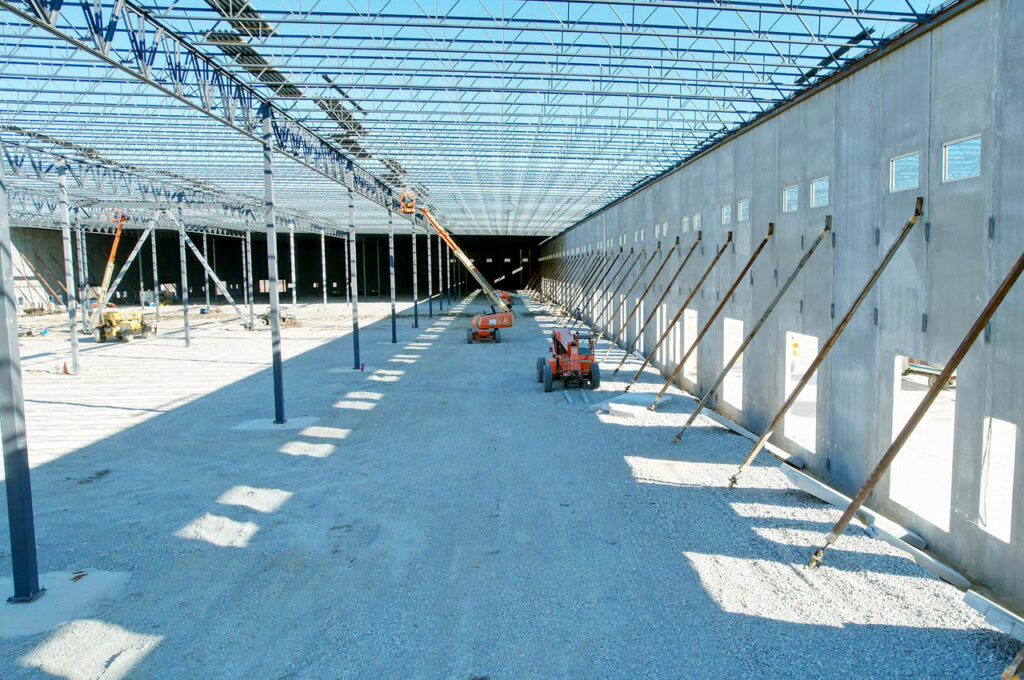 Wide interior view of a large industrial building under construction, featuring concrete walls, steel roof framing, and construction lifts on a gravel floor.