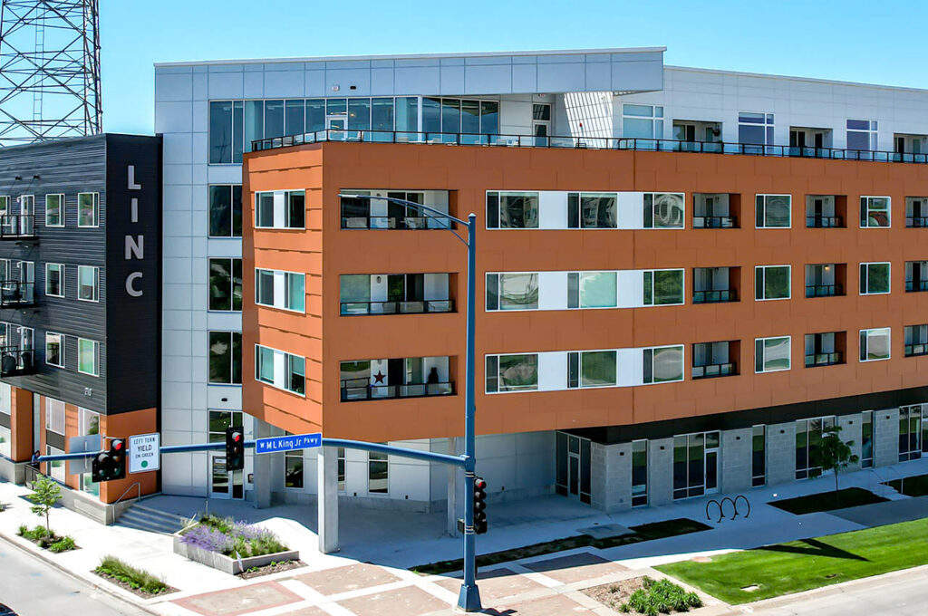 Corner view of a modern apartment building with orange and black exterior panels, large windows, and ground‑level commercial-style entrances.