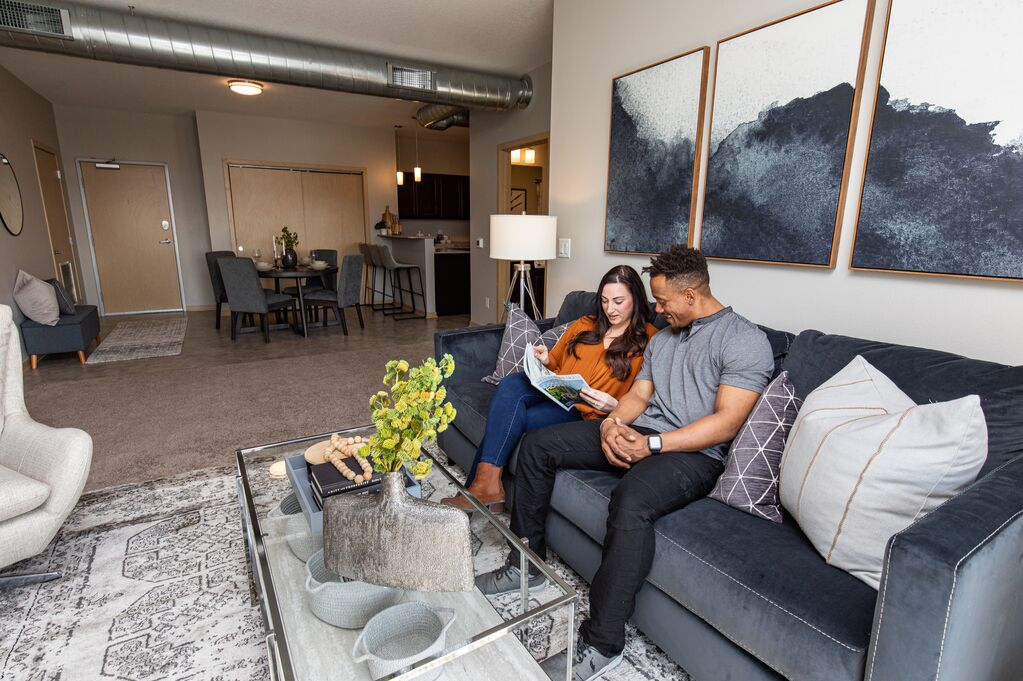 A couple sits together on a sofa in a modern apartment living room, reading a magazine at a glass coffee table with contemporary decor and artwork.