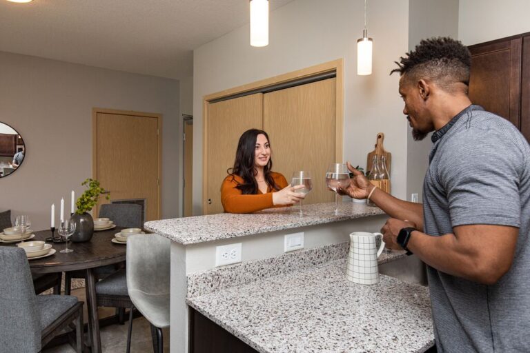 Two people clink glasses across a kitchen island in a modern apartment, with a dining area and contemporary lighting in the background.