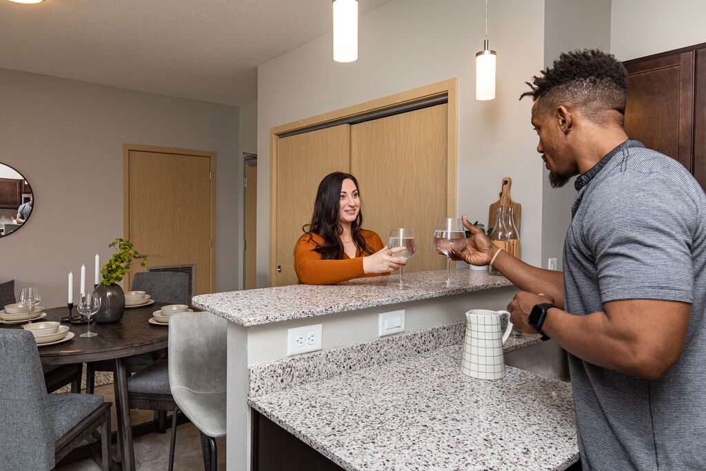 Two people clink glasses across a kitchen island in a modern apartment, with a dining area and contemporary lighting in the background.