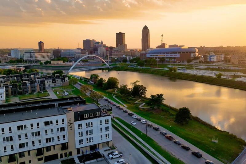 Aerial view of the Des Moines skyline at sunset, showing downtown buildings, a river with a pedestrian bridge, and residential apartments along the riverfront.