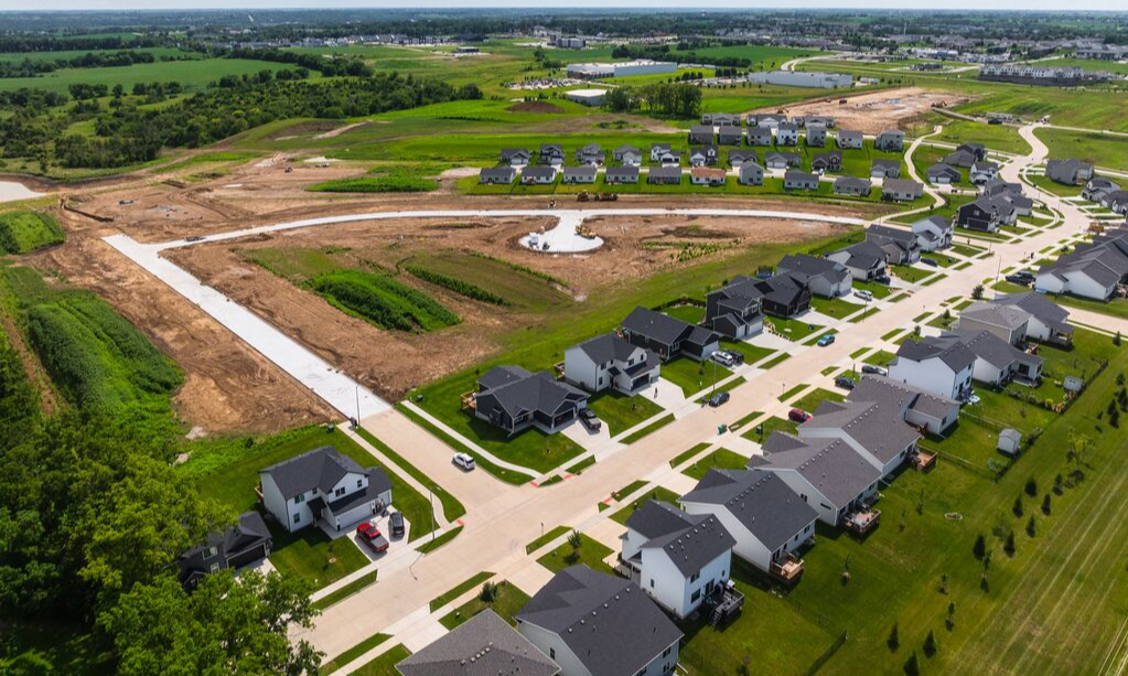 Aerial view of a growing residential neighborhood with newly built homes, paved streets, green lawns, and surrounding open land and farmland.