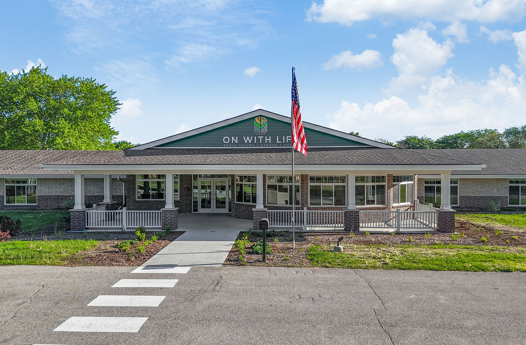Front exterior of the Hubbell‑built On With Life Long Term Skilled Care Facility with covered porch, brick façade, and a prominent flagpole.
