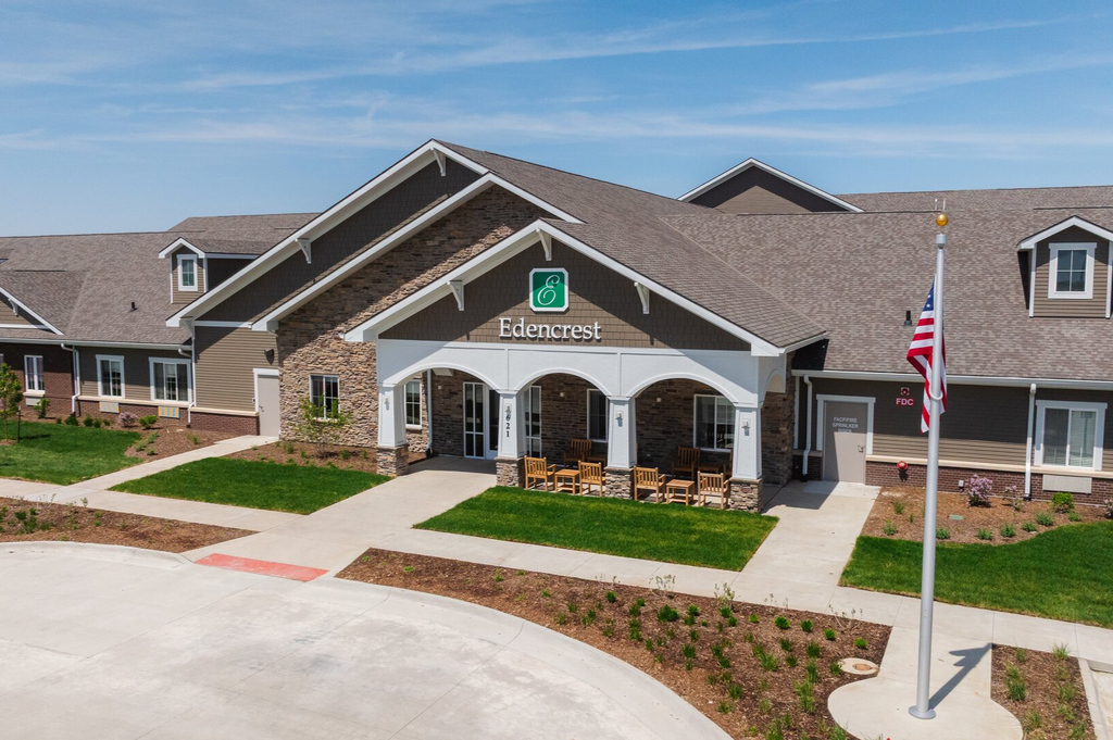 Aerial view of the Edencrest building featuring a covered entrance with stone columns, outdoor seating, landscaped grounds, and an American flag.